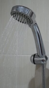 Vertical shot of a chrome shower head and hose mounted on a beige tiled wall with water running steadily.