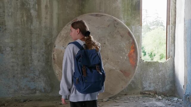 Person wearing backpack stands facing large circular concrete drainage pipe in deteriorated industrial building with natural light streaming through window, capturing solitude in decay