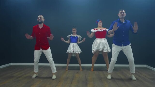 Man in blue shirt, woman in red dress with white collar, younger female in matching outfit perform bachata dance on wooden stage with teal backdrop, capturing coordinated movement