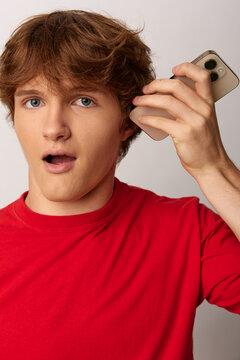 Young man in a red shirt reacts with surprise, holding a phone near his head. Studio shot with neutral background. Ideal for news, messages, or hype campaigns.