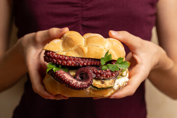 Italian sandwich made with tartaruga bread, grilled zucchini, octopus tentacles and parsley, held in the hands of a girl against a burgundy cloth. © Olga