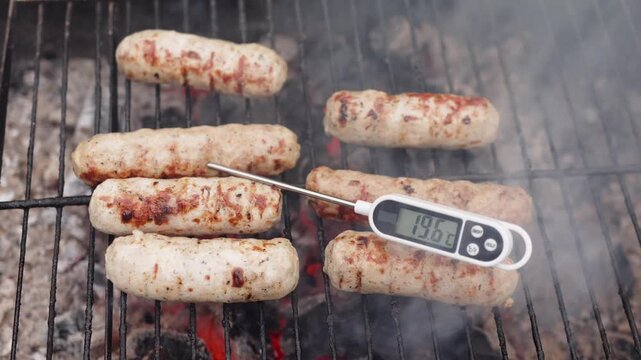 Checking internal temperature of roasting sausages with digital probe thermometer on outdoor barbecue grate. Smoke swirls around sizzling meat while electronic reader tracks rising heat.