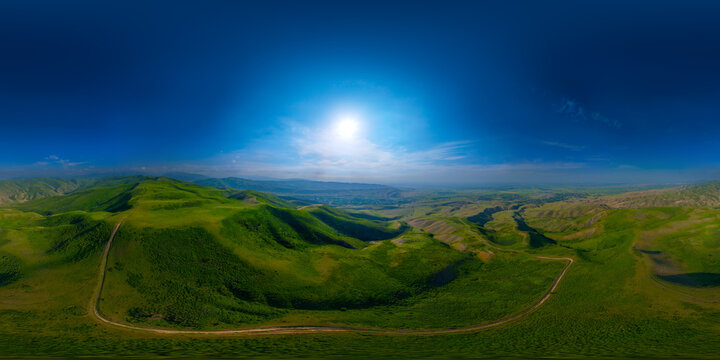 Full spherical panorama captured from aerial drone in Kyrgyzstan shows lush green hills and valleys. Bright sunlight illuminates the scene under a clear blue sky with wispy clouds.