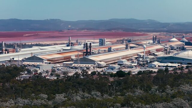 Aerial view of the industrial smelter facility with large factory buildings and red tailings ponds surrounded by forest near the coast in Boyne Island, QLD, Australia.