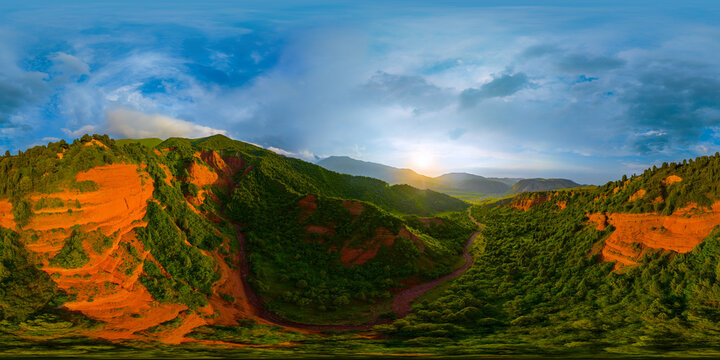 360-degree aerial panorama captures a mountain valley with lush green hills and steep orange cliffs. A winding dirt path runs through the valley floor under a blue sky with scattered clouds.