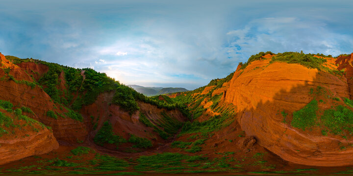 360-degree spherical panorama captures eroded clay hillsides and a deep ravine in Kyrgyzstan. Sparse green vegetation contrasts with reddish-orange soil under a partly cloudy sky at sunset.