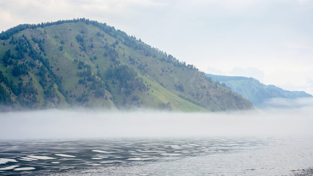 Green mountain rising from dark water with thick fog at the base. Overcast morning landscape of a misty Lake Baikal and forested hills. Nature of Siberia