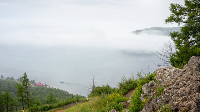High angle view of foggy Lake Baikal from a rocky mountain cliff, Siberia. Small boat navigating near a forested shoreline. Atmospheric nature landscape with copy space in background