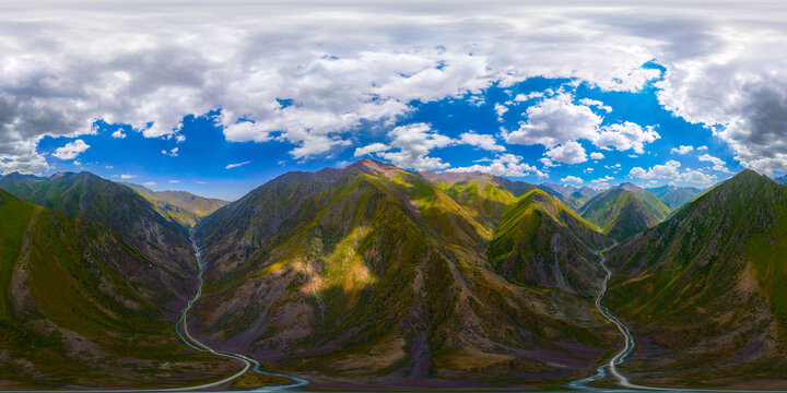 360-degree aerial panorama captures a rugged mountain landscape in Kyrgyzstan. Deep valleys, a winding road, and a narrow river are visible under a blue sky with scattered clouds.
