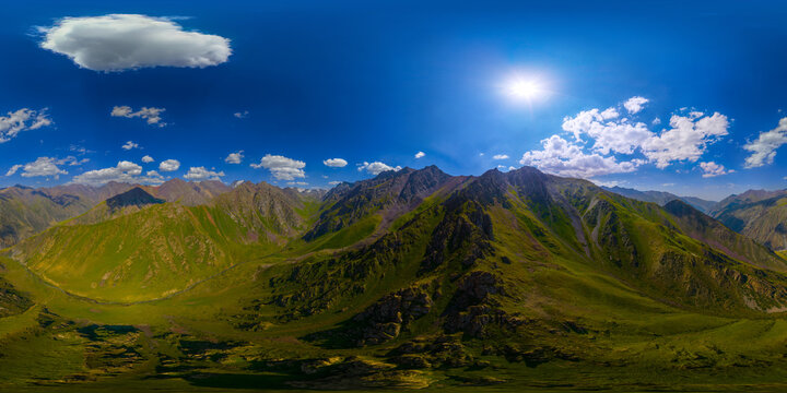 360-degree aerial panorama captures vast mountain landscape in Kyrgyzstan. Green grassy slopes, rocky peaks, and a winding valley with a stream are visible under a clear blue sky.
