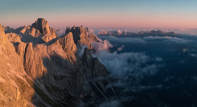 Aerial view of the Pale di San Martino peaks near Passo Rolle at sunrise, with warm alpenglow on rugged rock towers, drifting clouds and vast alpine mountain scenery in the Italian Dolomites. Colors: 