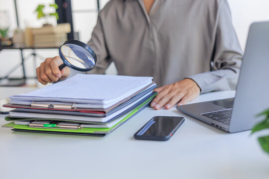 Professional closely inspecting a stack of organized documents and file folders on a desk with a magnifying glass, symbolizing a detailed financial audit, document review, or fraud investigation.