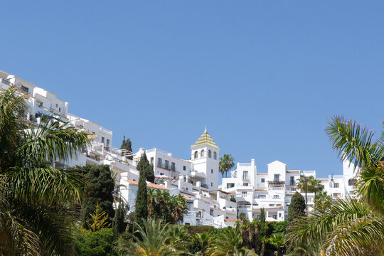 Nerja town showcasing typical white architecture built on a hillside, lush green vegetation, and palm trees under a clear blue sky, evoking a summer travel and holiday atmosphere