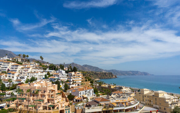 Nerja town displaying numerous white buildings and resorts along the hilly coastline, overlooking the blue Mediterranean Sea and distant mountains under a clear sky