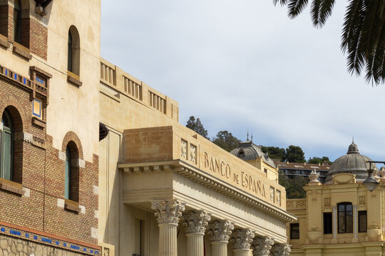 Architectural detail of the Banco de Espa&ntilde;a building in Malaga, showcasing its neoclassical design, columns, and intricate stone carvings under a partly cloudy sky