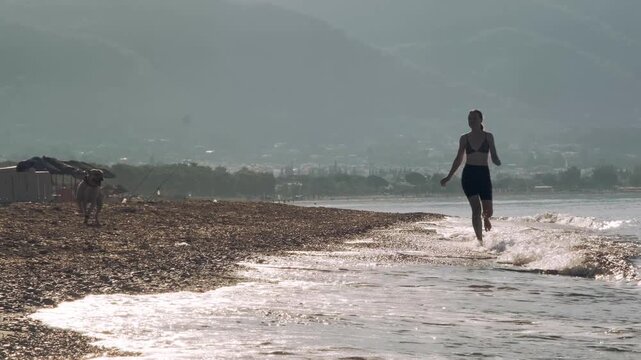 A video of an eight year old female one eyed pitbull running together with her thirty year old female owner on a sunny morning sandy beach in warm golden sunlight.