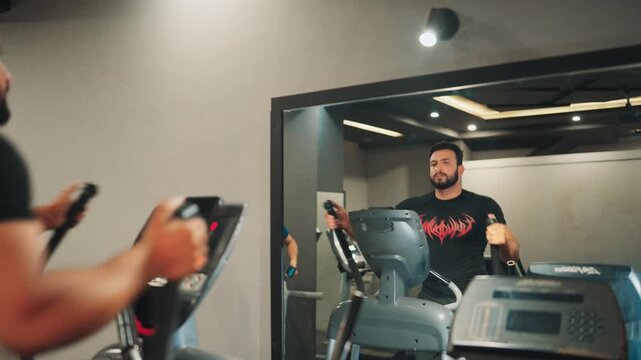 Over the shoulder mirror reflection shot of an athletic bearded man exercising on an elliptical cross trainer during an indoor cardio workout in a modern gym.