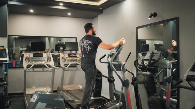 Wide side view video shot of an athletic bearded man exercising on an elliptical cross trainer for cardiovascular conditioning in a modern gym setting.