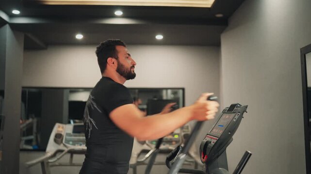 Medium side profile video shot of an athletic bearded man training on an elliptical cross trainer for cardiovascular endurance inside a modern gym facility.