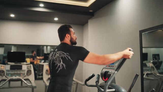Muscular male athlete in a black compression shirt operating an elliptical trainer machine during an active cardio workout routine in a fitness center.