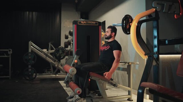 Wide shot of an athletic bearded man performing leg extension machine exercises for quadriceps hypertrophy and leg strength training in a gym setting.