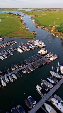Vertical aerial drone video of marina harbor, yachts, boats, river houseboats, windmill, green polder fields, and lakes in Warmond, South Holland, Netherlands at golden hour sunset