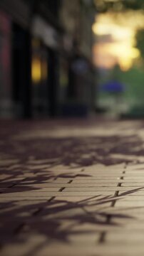 The scene shows a walkway during dusk with light reflections on a wet surface. Buildings are present in the background. The atmosphere suggests the end of the day.