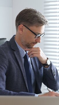 Adult businessman wearing blue suit and tail is concentrating and thinking while working at the desk in a bright, modern office, demonstrating professionalism and efficiency. Business people concept