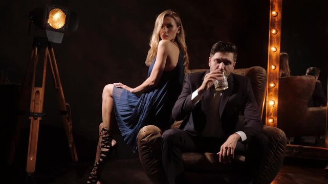 Noir backstage lounge with tuxedo man and woman in blue dress reclining on tufted sofa