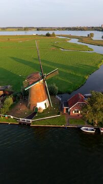 Vertical aerial drone video of traditional Dutch windmill, river canal, green polder fields, and lake landscape in Warmond near Leiden, South Holland, Netherlands at golden hour sunset
