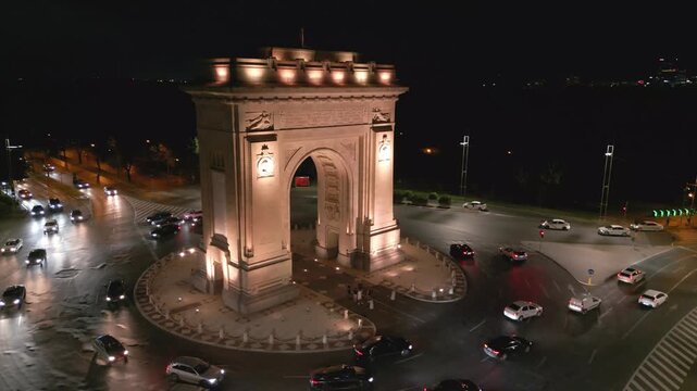 Nighttime aerial drone shot of the illuminated Arch of Triumph in Bucharest, Romania. View of the monument with city lights, traffic light trails, and evening urban atmosphere.