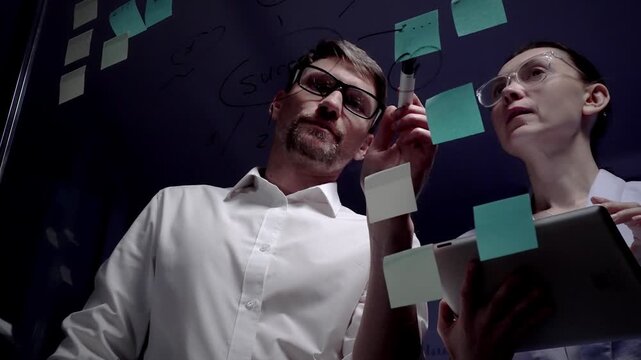 Business colleagues collaborating on new innovative project, drawing mind map with success word on clear glass writing board with sticky notes in modern office environment