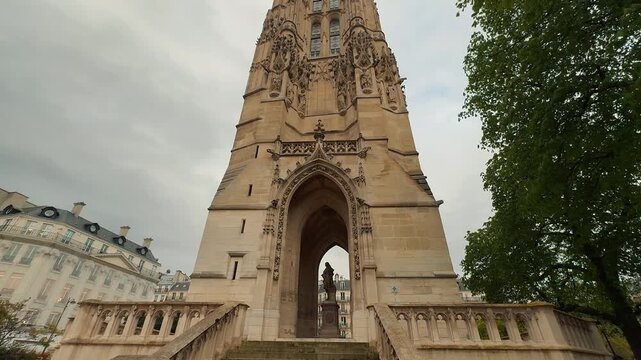 A smooth tilt shot revealing the intricate Flamboyant Gothic architecture and ornate carved stonework of the historic Tour Saint Jacques against an overcast sky in central Paris, France.

