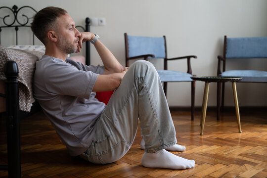 Lonely depressed man sits on floor beside bed at home looking withdrawn and hopeless while struggling with sadness, isolation and personal crisis.