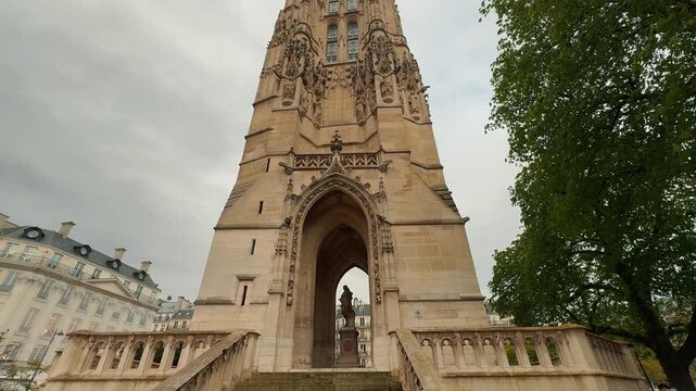 A smooth tilt shot revealing the intricate Flamboyant Gothic architecture and ornate carved stonework of the historic Tour Saint Jacques against an overcast sky in central Paris, France.

