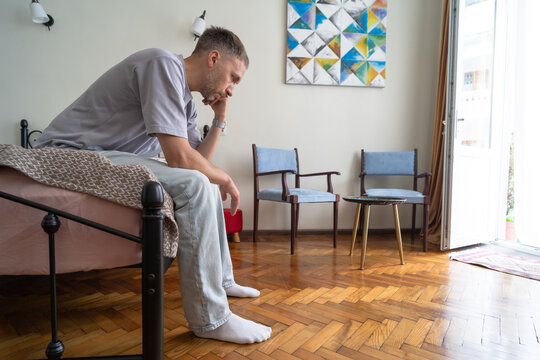 Exhausted man sits on edge of bed at home feeling stressed and overwhelmed while thinking about personal problems, uncertainty and difficult life decision.