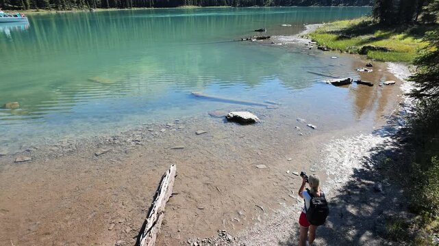Woman standing on shore of Spirit Island in Maligne Lake, holding camera and capturing Spirit Island with its clear water reflecting surrounding forest in Jasper National Park in Alberta of Canada