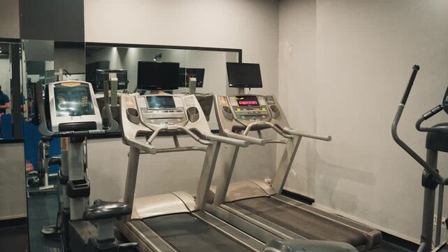 Wide view video shot of rowed electric treadmills with display screens positioned in front of a mirror inside an empty commercial gym