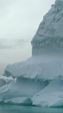 Vertical Video of Angular Iceberg With Snowy Mountains Behind. White ice, rippling water and snow-covered ridges create a layered cold seascape beneath soft diffuse light.