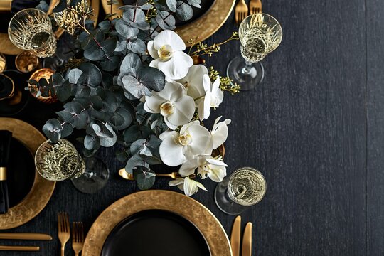 Elegant table setting with orchids and eucalyptus leaves. Luxurious gold chargers adorn black plates for a festive meal. Sophisticated dining ambiance with sparkling champagne