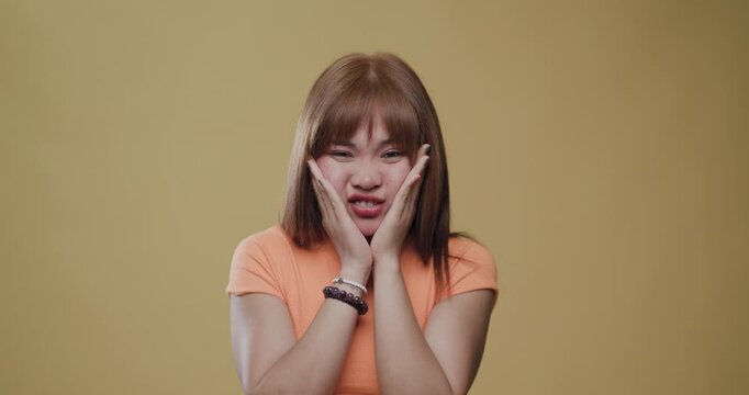 Young Asian woman making a disgusted face with hands on cheeks against a yellow background