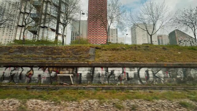 Scenic view from a moving train window passing urban graffiti walls, modern skyscraper towers, and waiting commuters along the platform of Javel station in Paris, France, during daytime.

