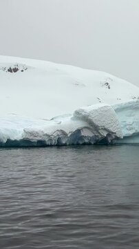 Vertical Video of Snowy Iceberg Ridge Across Calm Water. White ice, rippling water and snow-covered ridges create a layered cold seascape beneath soft diffuse light.