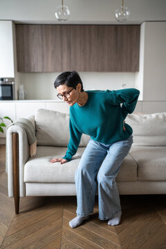 Senior woman with spinal disorder suffers from sudden back pain after rising from sofa. Elderly female struggling with muscles spasm in lower back, chronic herniated discs and protrusions at older age