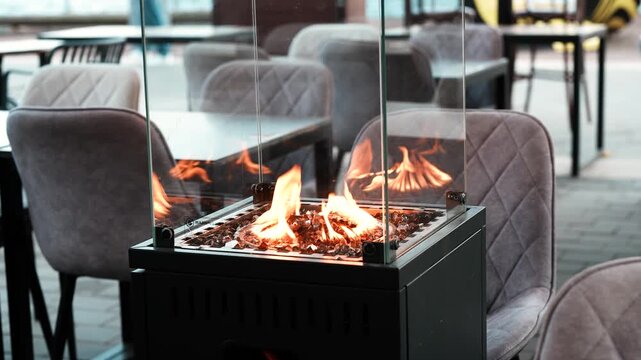 Outdoor terrace in restaurant with glass gas heater for guests comfort and relaxation, open-air patio in dining area with comfortable chairs and tables beside gas fireplace in glass box.