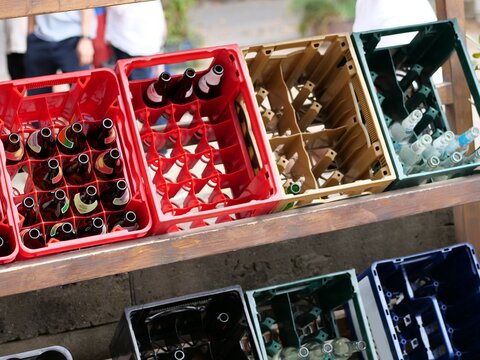 Rows of red, yellow, and green plastic drink crates containing glass beer and soda bottles stored on a wooden shelf frame at an outdoor festival.