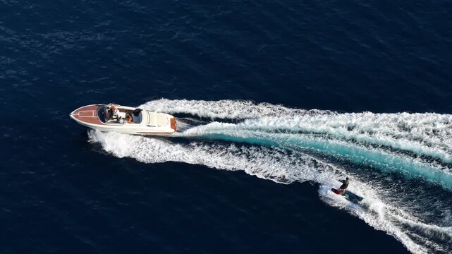 Aerial view of a motorboat towing a person on a wakeboard through the deep blue sea near the Le Trayas coastline in Saint-Raphael, Provence-Alpes-Cote d'Azur, France.