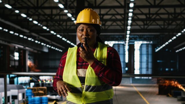 Black female engineer uses a walkie-talkie on a production line, supporting portable communication and heavy industry operations in a machinery hall.