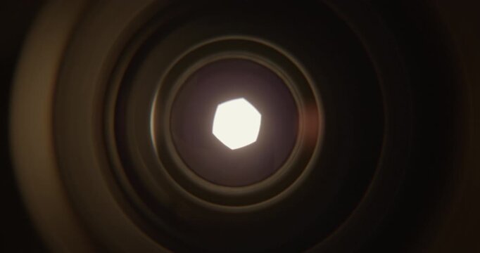 Extreme closeup of a camera lens aperture slowly opening as intense bright light emerges from behind the blades. Cinematic visual representing photography, optics, focus, exposure, illumination.