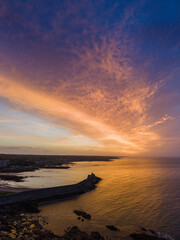 Fototapeta premium Aerial view of coastal pier at golden sunset. Warm light reflects on water and stone breakwater. Calm sea meets land with sandy beach nearby. Spiddal, Ireland, Galway bay. Travel and tourism.